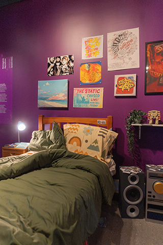 The view of a warmly lit bedroom. A twin sized bed with a green duvet is seen at the center of the image, and in large feature are the paintings on the purple wall above the bed, a bright bedside light, and what appears to be a stereo speaker on the floor.