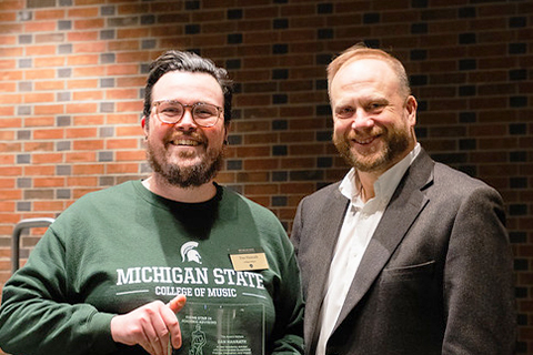 A person wearing a dark green sweatshirt with the words ‘Michigan State College of Music’ printed on the front stands indoors holding a clear glass award plaque with both hands. Another person stands beside them, wearing a dark blazer and light shirt. Both are positioned in front of a background made of dark red brick.