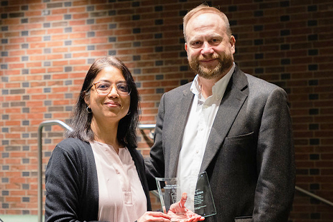 A person wearing glasses, a light-colored blouse, and a dark cardigan holds a clear award plaque with both hands. Another person stands to the right wearing a blazer and light shirt. They are positioned in front of a brick wall in an indoor auditorium-style setting. The lighting is focused on the two individuals.