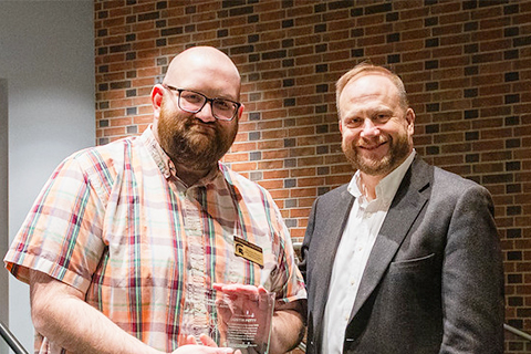 A person wearing a short‑sleeve plaid shirt holds a transparent award plaque with both hands while standing in an auditorium-style space. Another person stands next to them wearing a blazer and light shirt. The background features a dark red brick wall, a metal handrail, and steps leading upward.