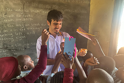 A person standing in a classroom, holding papers while several young students gather around and reach toward the materials. A chalkboard behind them displays handwritten sentences about brushing teeth.