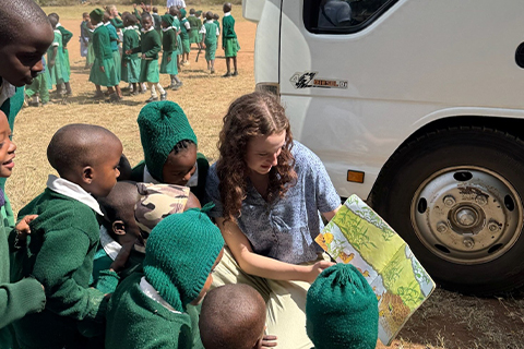 A person sitting on the ground next to a vehicle, holding an illustrated children’s book open while several students in green uniforms gather around to look at the pages. More students stand in the background on a grassy field.