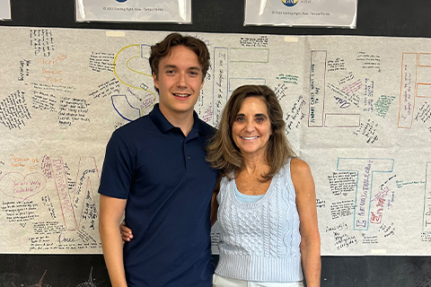 Two people standing in front of a large poster covered with handwritten messages and signatures. The poster features large outlined letters filled with notes.