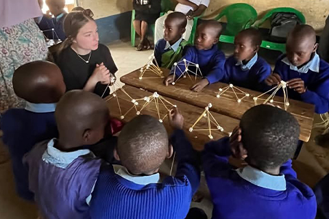 A person seated at a wooden table with a group of students wearing matching uniforms. The students are building small structures made of sticks and marshmallows or similar connectors.