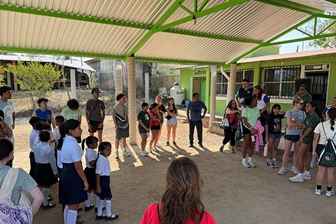 Large group of people standing in a circle under a shaded structure in an outdoor area. A mix of school-age children and older individuals are present, with buildings and trees in the background.