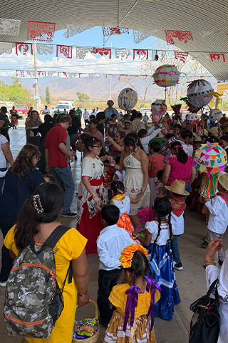 Indoor gathering with many people participating in a cultural celebration. Individuals wear brightly colored traditional-style clothing, with large decorated paper ornaments hanging overhead.