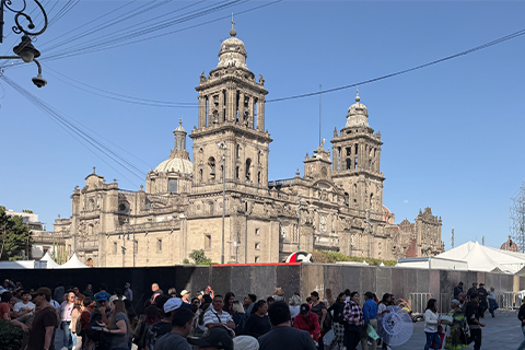 Exterior view of a large historic cathedral with ornate stone towers and architectural details. A crowd of people stands in the foreground, with tents and fencing visible near the building.