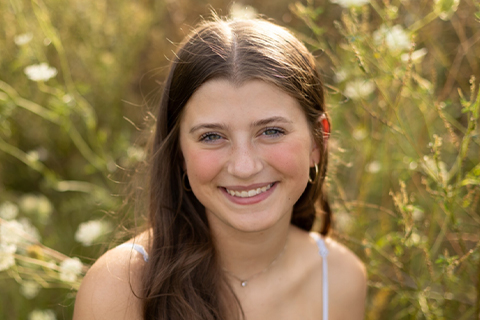 Outdoor portrait of a scholarship recipient smiling toward the camera, standing among tall green plants. The person is centered in the frame with a softly blurred natural background.