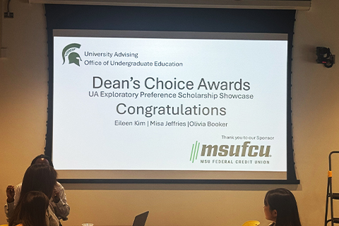 Projected slide reading ‘Dean’s Choice Awards – UA Exploratory Preference Scholarship Showcase. Congratulations Eileen Kim and Misa Jeffries and Olivia Booker.’ University Advising, Office of Undergraduate Education branding appears at the top, with MSU Federal Credit Union listed as sponsor. Audience members are visible seated in front of the screen.