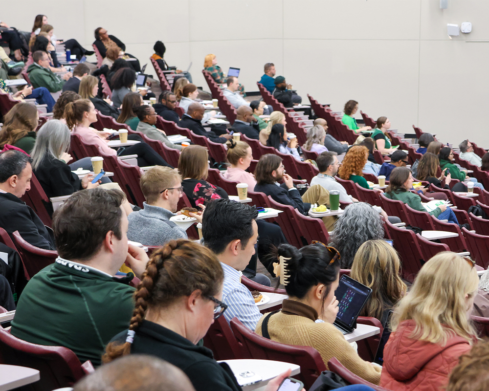 Participants at a campus event sit in tiered lecture hall seating, viewing a presentation and taking notes on devices and notepads.