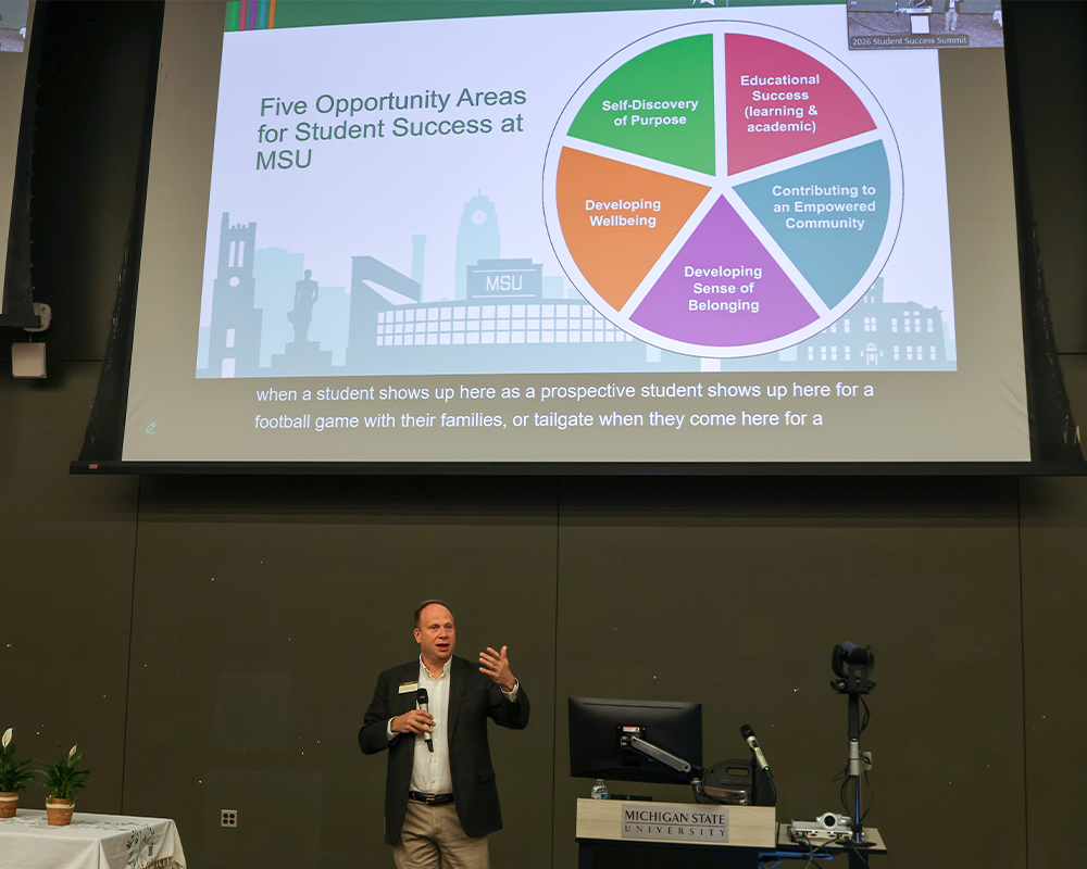 Presenter stands at the front of a lecture space while a projected slide titled “Five Opportunity Areas for Student Success at MSU” displays a circular diagram with categories such as self-discovery of purpose, developing wellbeing, sense of belonging, community contribution, and educational success.