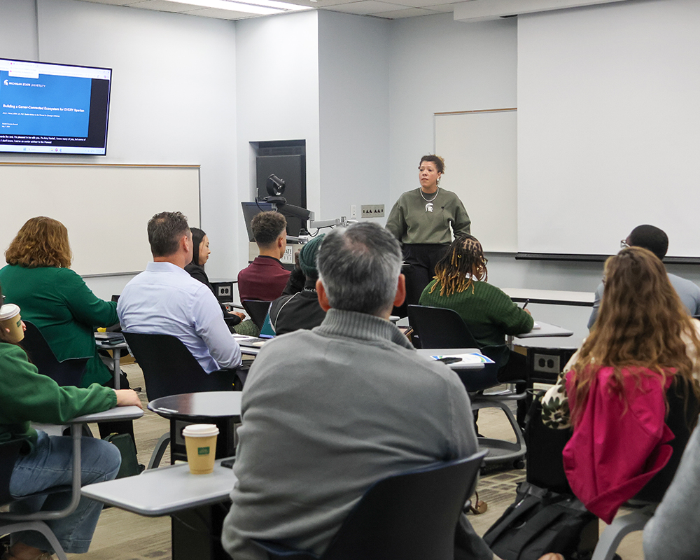 Facilitator leads a discussion in a classroom setting as participants sit in small desk groups, listening and viewing a screen presentation.