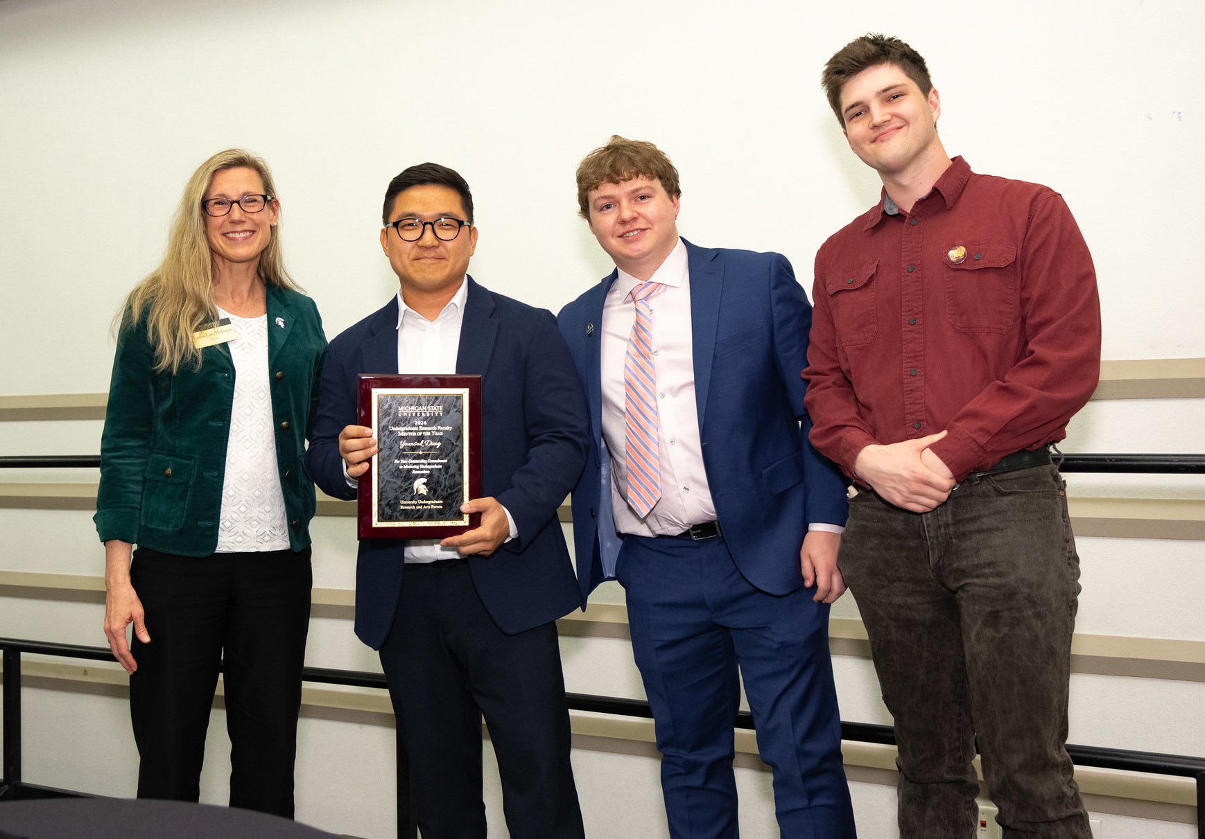 Four individuals pose for a recognition photo indoors, with one person holding a Michigan State University award plaque.