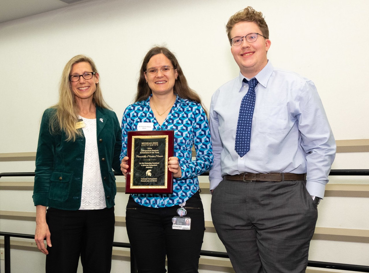 Three individuals pose with a Michigan State University award plaque during a recognition event, with the award recipient standing in the center holding the plaque.