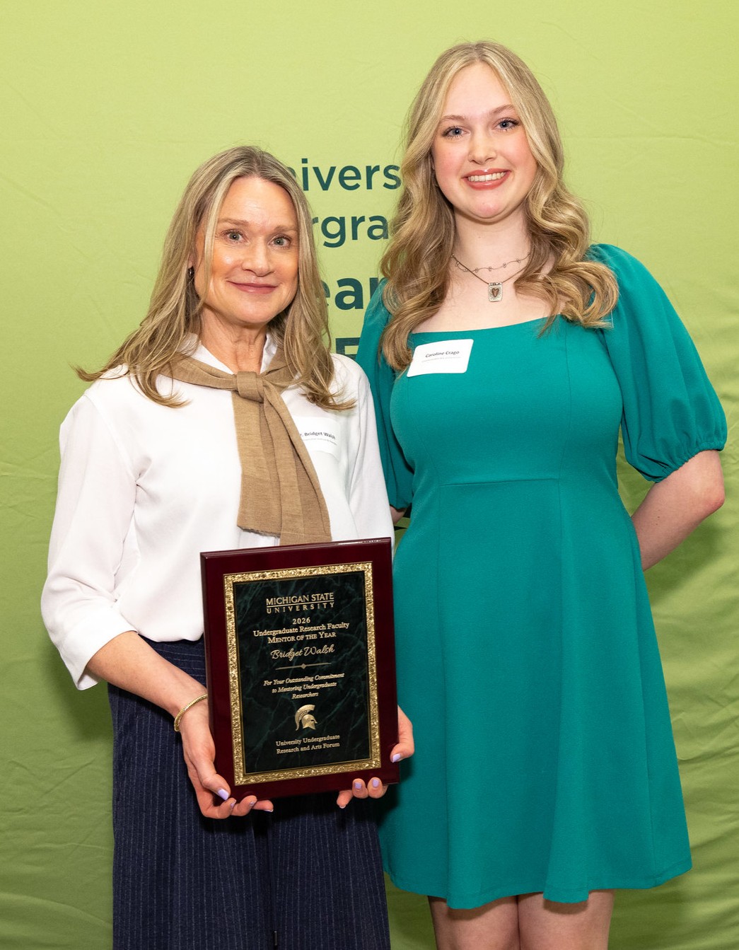 Two individuals pose in front of a University Undergraduate Research and Arts Forum backdrop, with one holding a Michigan State University Mentor of the Year award plaque.