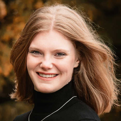 Professional headshot of a person with shoulder-length hair wearing a black turtleneck, photographed outdoors with a blurred autumn background.