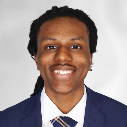 Professional headshot of a person wearing a blue suit jacket, white shirt, and patterned tie against a light gray background.