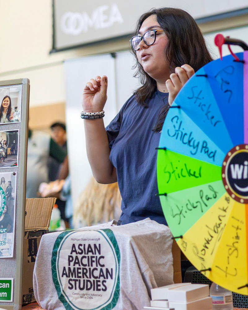 Student representative speaks at an Asian Pacific American Studies table beside a colorful prize wheel at a campus involvement fair.