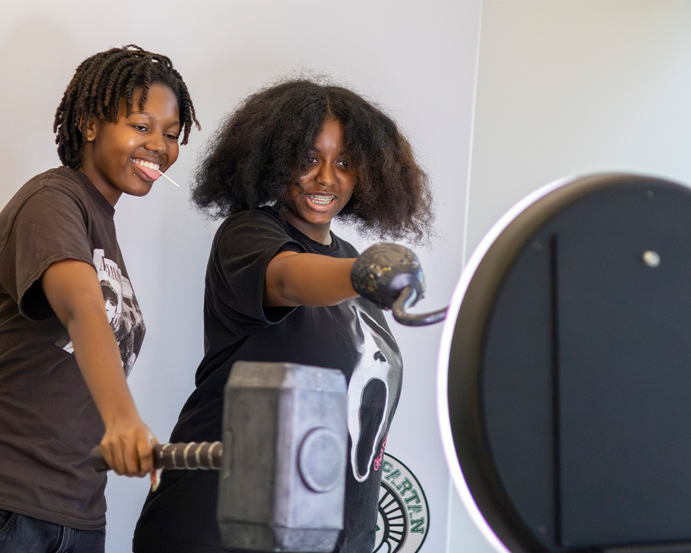 Students pose with oversized prop hammers beside a ring light and camera setup at a campus event activity booth.