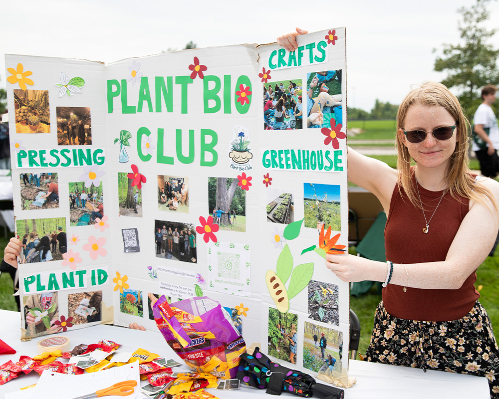 Student stands beside a Plant Bio Club display board featuring photos, plant-themed crafts, and information about club activities at an outdoor student organization fair.