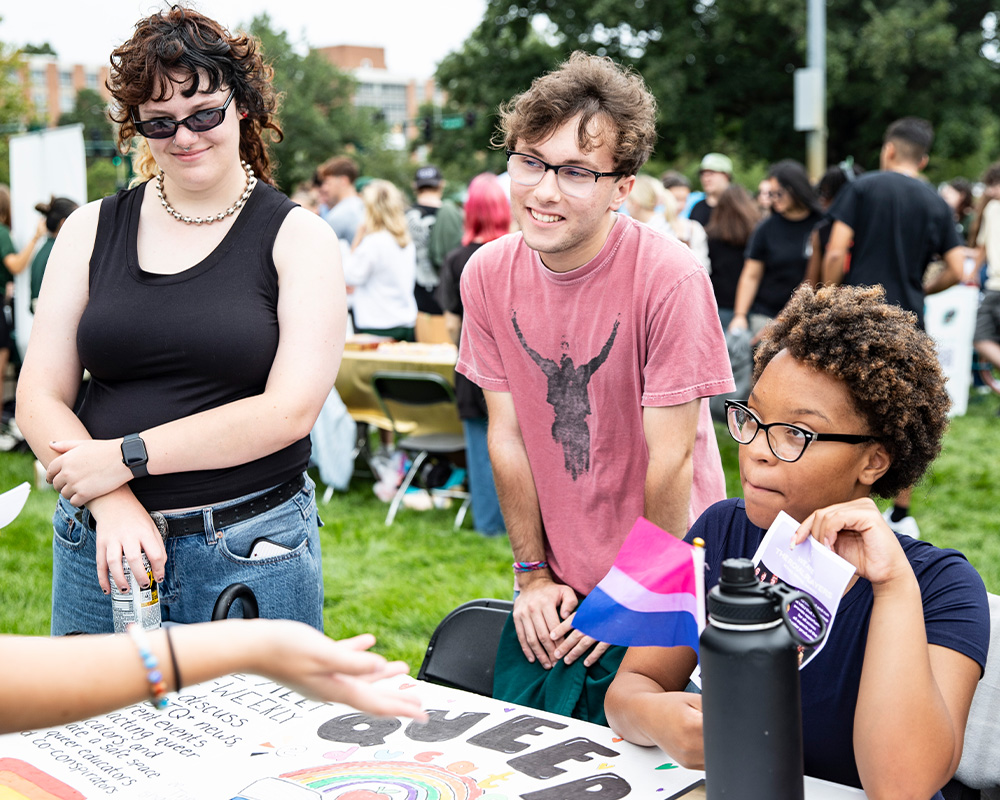 Students gather around a student organization table, discussing information displayed on a poster during an outdoor campus involvement event.