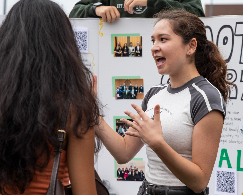 Student speaks with a visitor in front of a student organization display board during an outdoor campus involvement fair.