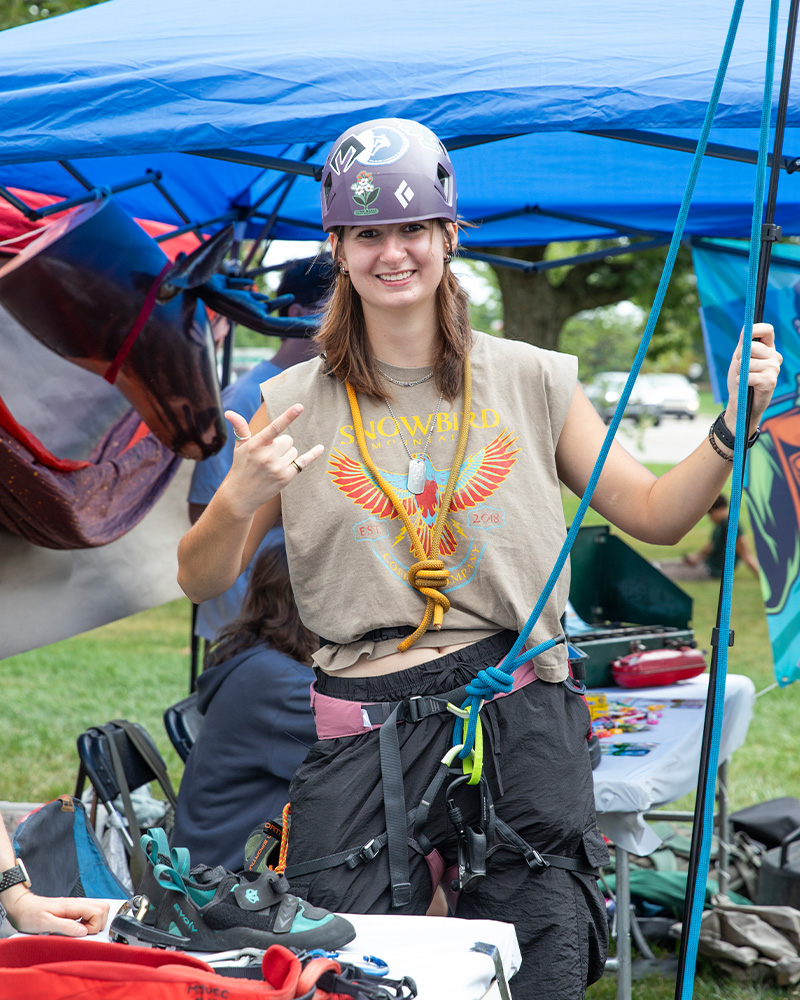 Student wearing a climbing helmet, harness, and ropes poses at an outdoor recreation organization booth during a campus involvement fair.