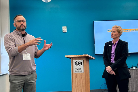 Two conference participants stand at the front of a meeting room during a presentation, with one speaking and gesturing beside a podium.
