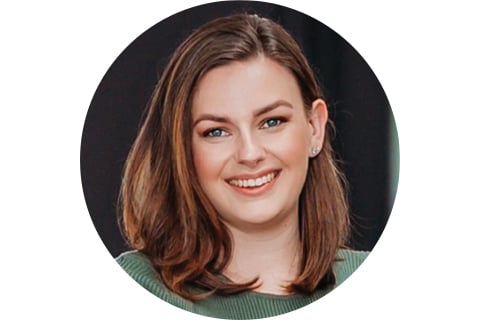 Portrait in a circle frame. Subject is set against a black background, viewed from the shoulders up, looking at the camera and smiling. They have shoulder length brown hair pushed to one side and are wearing a green sweater.