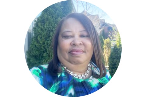 Portrait. Subject stands in front of green foliage while looking at the camera. Their hair is long at their shoulders and are wearing a greenish-blue abstract shirt. 