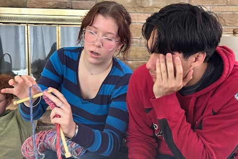 Two people sit close together indoors as one demonstrates how to knit using wooden needles and multicolored yarn.