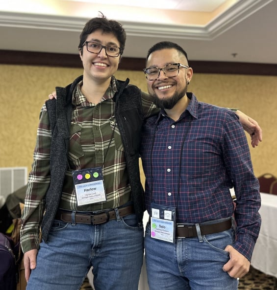 Two people stand next to one another smiling at the camera. They have their arms over each other's shoulders and are wearing nametags on a lanyard. 
