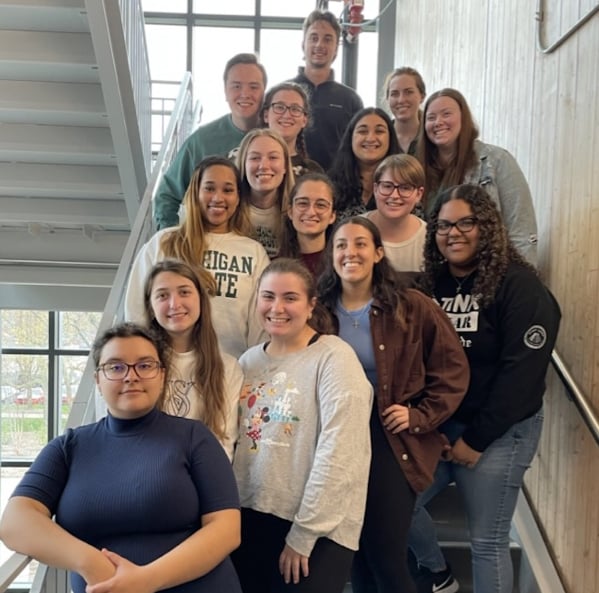 A group of students stands on a staircase looking at the camera and smiling. They are situated on ascending steps, wearing various clothing items, but primarily jeans and comfortable looking sweatshirts and shirts.