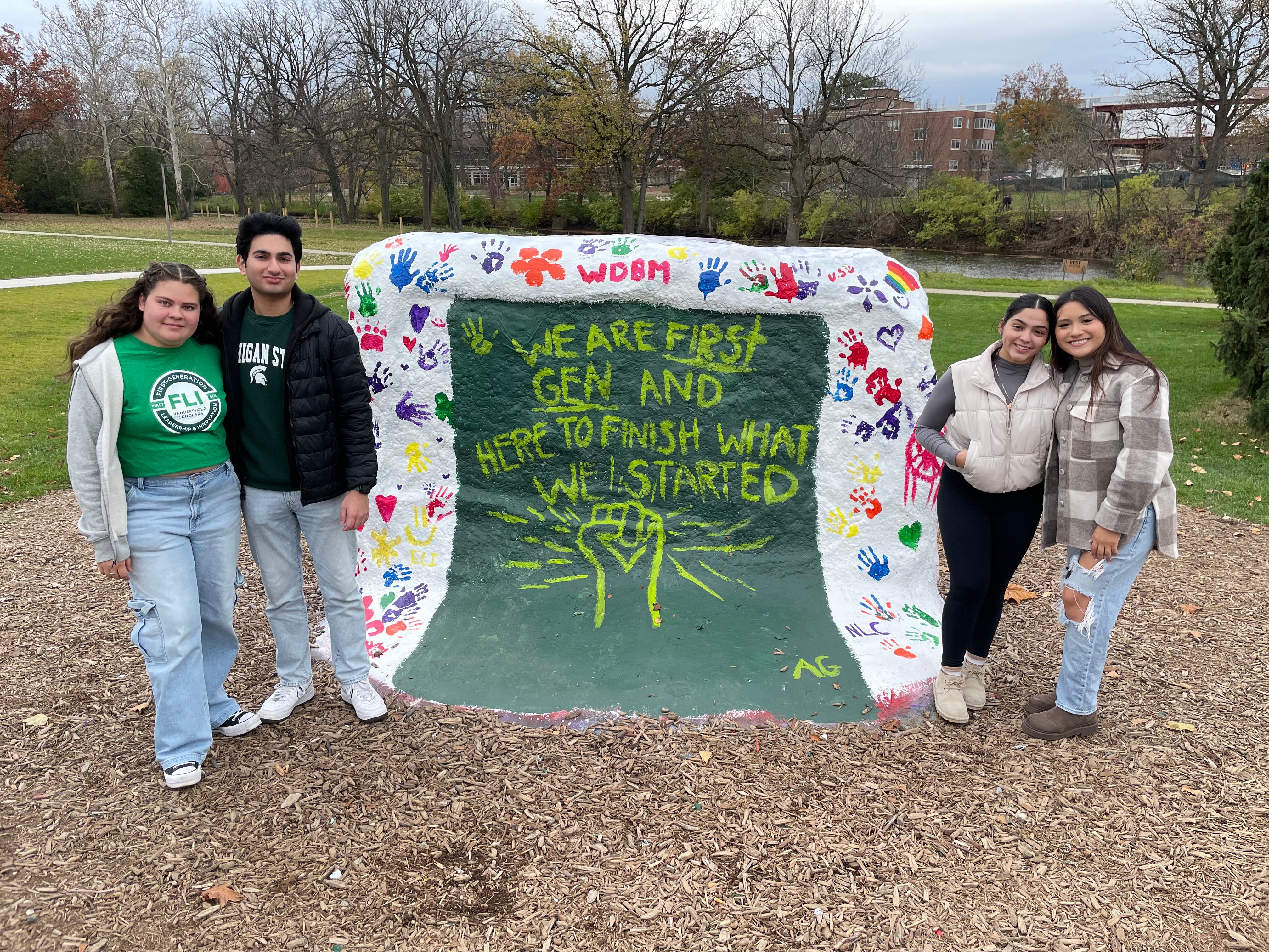 Four FLI students stand around The Rock on MSU's campus which is painted white with colorful handprints and words reading We are First Gen and here to finish what we started!