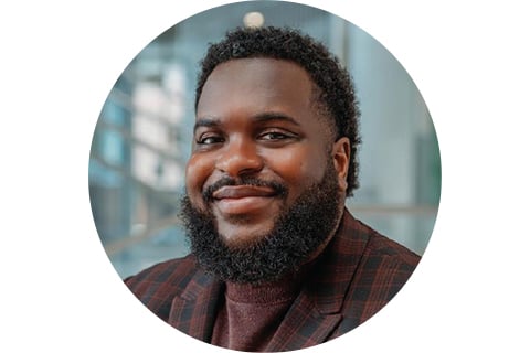 Portrait in a circle frame. Subject is looking at the camera with a smile, seen from the shoulders up. They are wearing a brown coordinated suit jacket and shirt, with a black beard and short black hair.