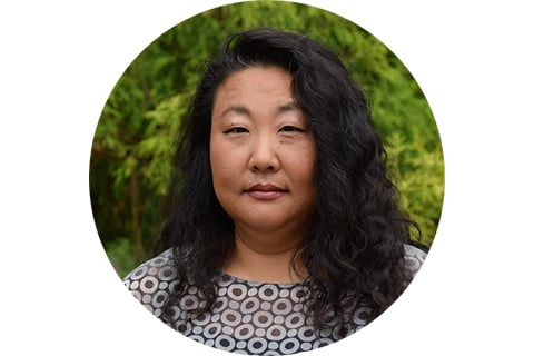Portrait. Subject is looking at the camera and standing in front of green foliage, with their long dark hair at their shoulders. They are wearing a black and white shirt with geometric designs.