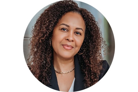 Portrait in a circle frame. Subject is facing toward the camera seen from the shoulders up, with a small smile. They have long, full curly hair, with a dark suit jacket and necklace on. 