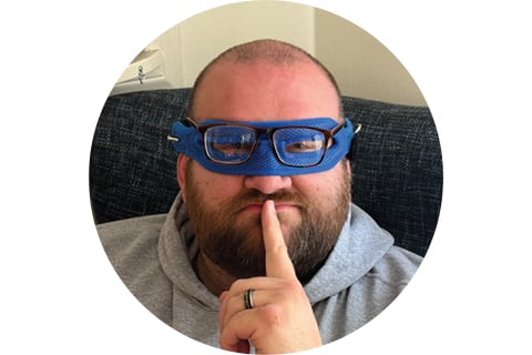 Portrait in a circle frame. Subject is seen from the shoulders up, looking at the camera with their finger over their mouth in a pensive manner. They are wearing a blue superhero partial mask over their eyes.