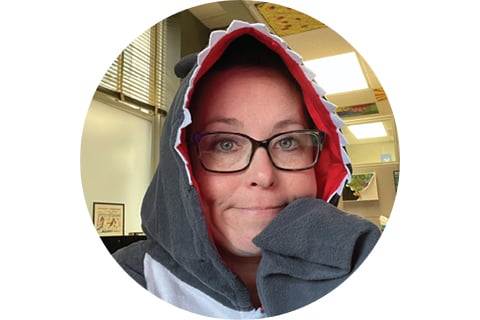 Portrait in a circle frame. Subject is looking at the camera and is wearing a subtle shark costume, with the hood over their head lined in shark "teeth." They are wearing glasses and have one arm (fin) resting on their chin. 