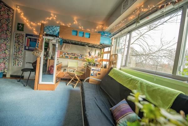 View of a dorm room. There is a large window on the right with a couch under it, with a desk and mirror next to it, under a lofted bed.