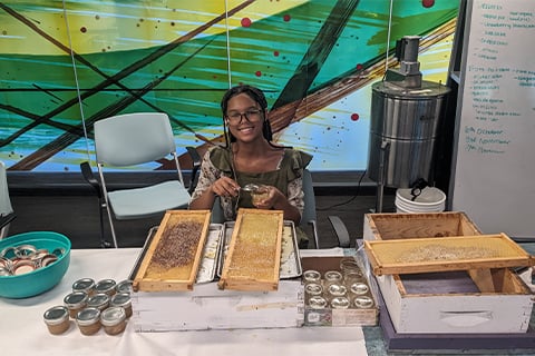 A student sits at a table with two honeycomb sections from a bees nest in  front of them. Next to them are jars of honey and other honey products.