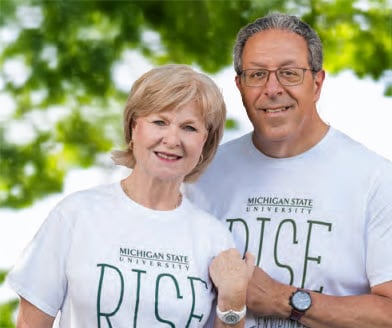 Two people stand side-by-side looking at the camera. They are both wearing white shirts that say Michigan State University and RISE on the front. The are standing in front of a blurry green and white colored background.