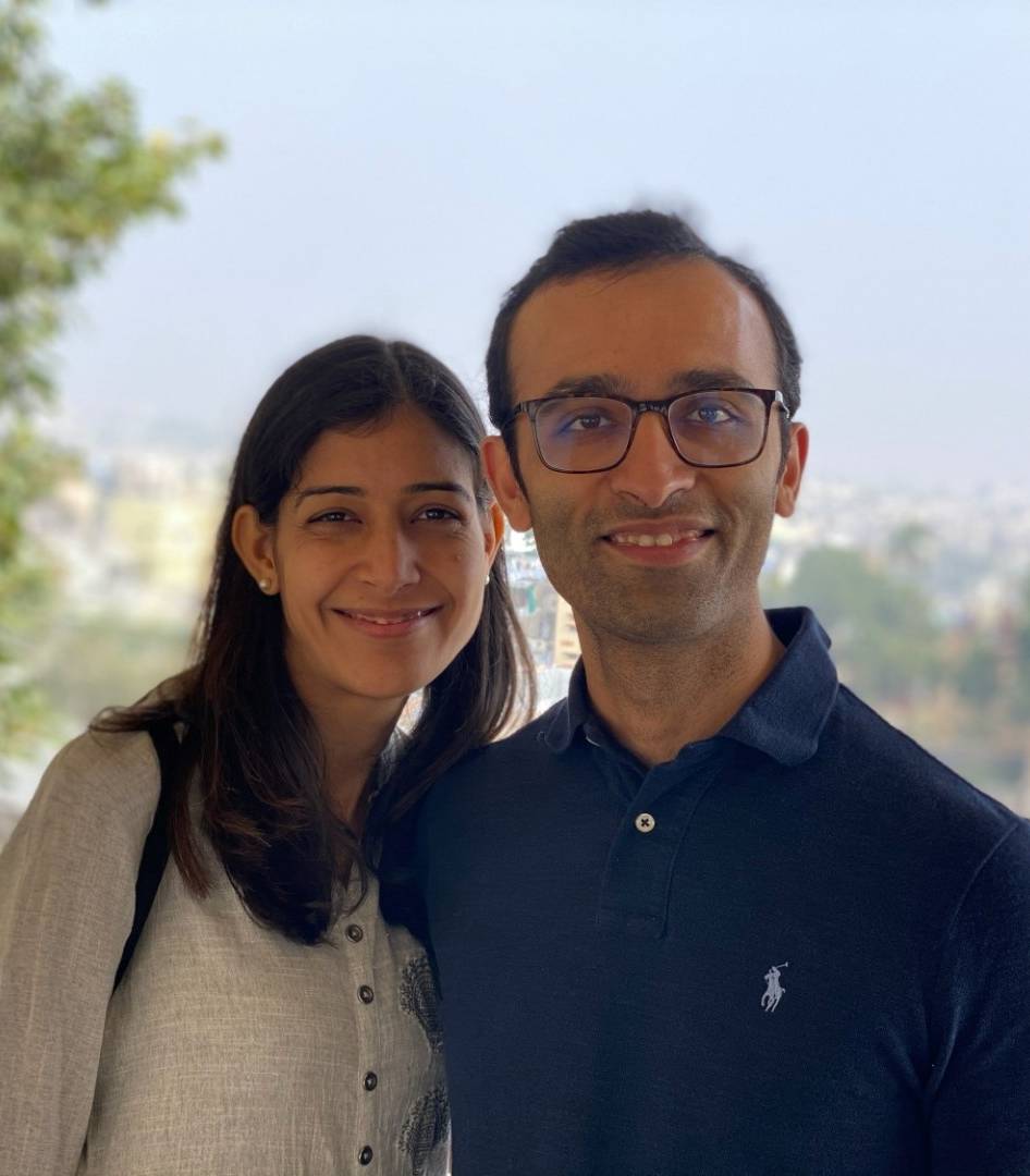Two people stand side by side looking at the camera and smiling. The person on the right is taller with short dark hair and glasses, and the person the right has long dark hair and is wearing a white cardigan. The view behind them is of a distant cityscape.