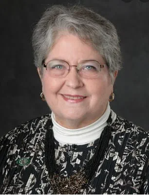 Portrait. The person looks at the camera while smiling, wearing a black and white jacket watercolor style jacket, with a white turtle neck. Their hair is short and grey, and they are wearing glasses. 