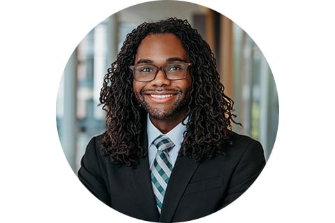 Portrait. Subject is wearing a dark suit jacket, with a light blue collared shirt, with a similarly colored striped tie. They are looking at the camera and smiling while wearing glasses with dark curly locks down to their shoulders.