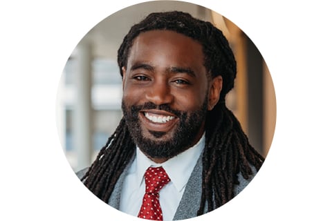 Portrait in a circle frame. Subject is seen from the shoulders up looking at the camera and smiling. They have long locks down the sides of their shoulders and are wearing grey suit jacket with a bright red tie and white dress shirt.
