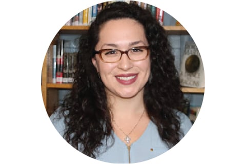 Portrait in a circle frame. Subject is looking at the camera and smiling and is seen from the shoulders up. They have long, dark hair and are wearing glasses, standing in front of a bookshelf.
