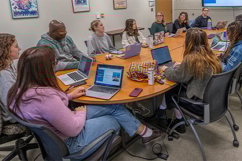 A classroom group sits around a brown oval table with a large screen at one end of the table. Students have books, laptops and other classroom materials spread out on the table.