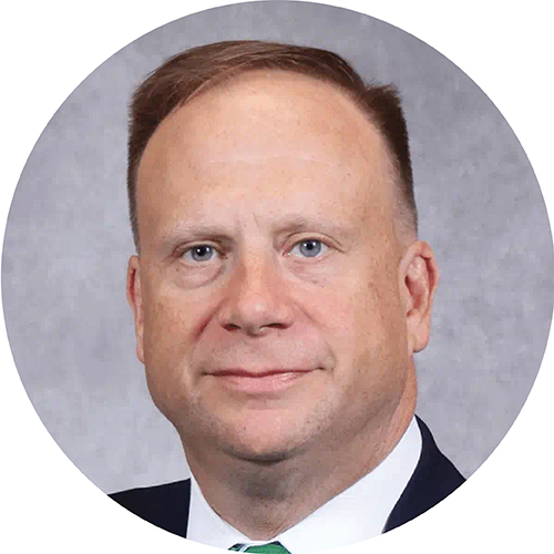 Portrait framed in circle. Subject is looking at the camera and can be seen from the shoulders up, while wearing a suit and tie with short, dark hair. They are standing against a grey backdrop. 
