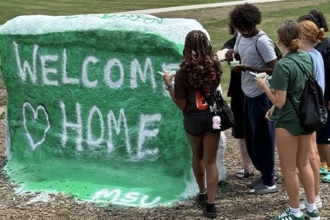 A small group of students stand to the right of MSU's rock which is painted green with white words reading "Welcome Home" with a heart shape.